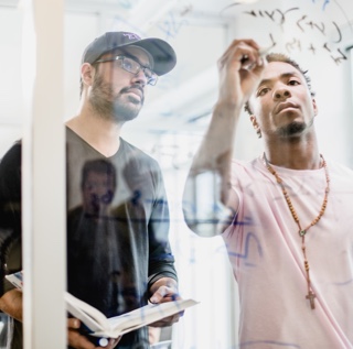 One male student holding a book looks at what another male student is writing on a glass wall