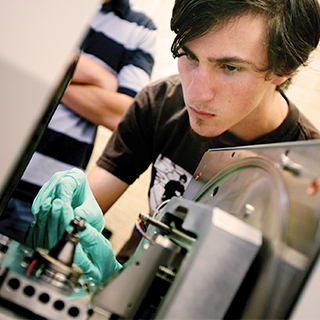 A male student wearing green protective gloves works on a piece of electronics