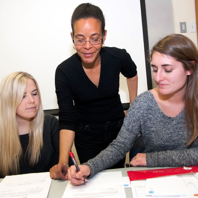 An economics professors looks on as one of two students writes out an idea on a piece of paper