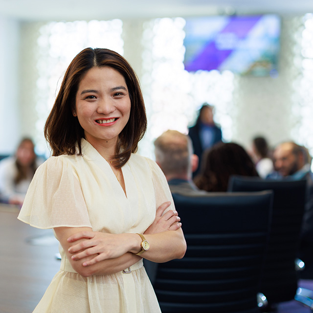 MBA student stands, smiling, while others gather around the boardroom table