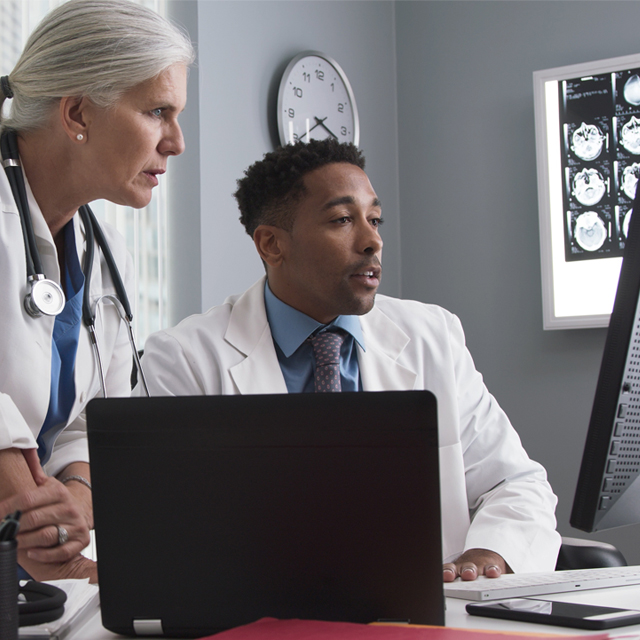 Two medical professionals in white coats, one sitting at a desk, the other looking over his shoulder at a computer monitor.