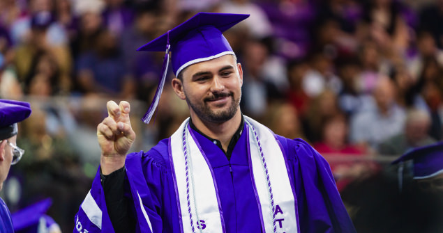 Student athlete, dressed in full graduation regalia, gives the Go Frogs hand sign as he walks across the stage